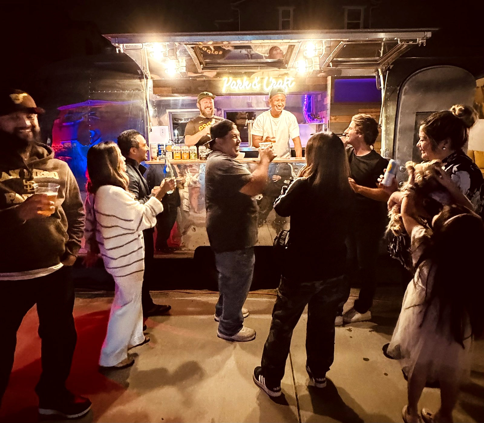 Guests gathered around the Park & Craft Airstream bar at night with Chris and Mark serving